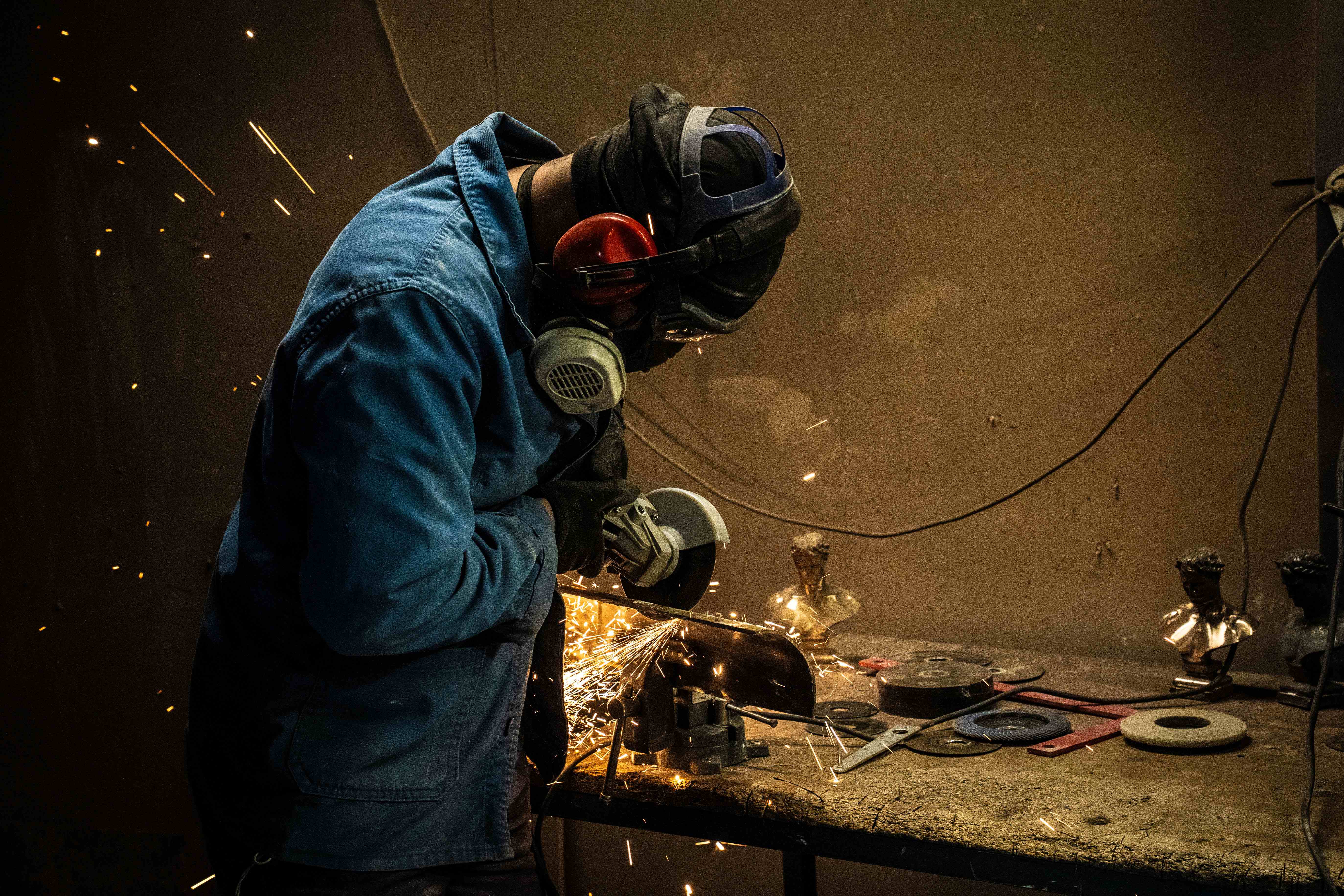 Protégé aux au visage et aux oreilles, un artisan utilise sa meuleuse pour poncer une statuette en bronze. 