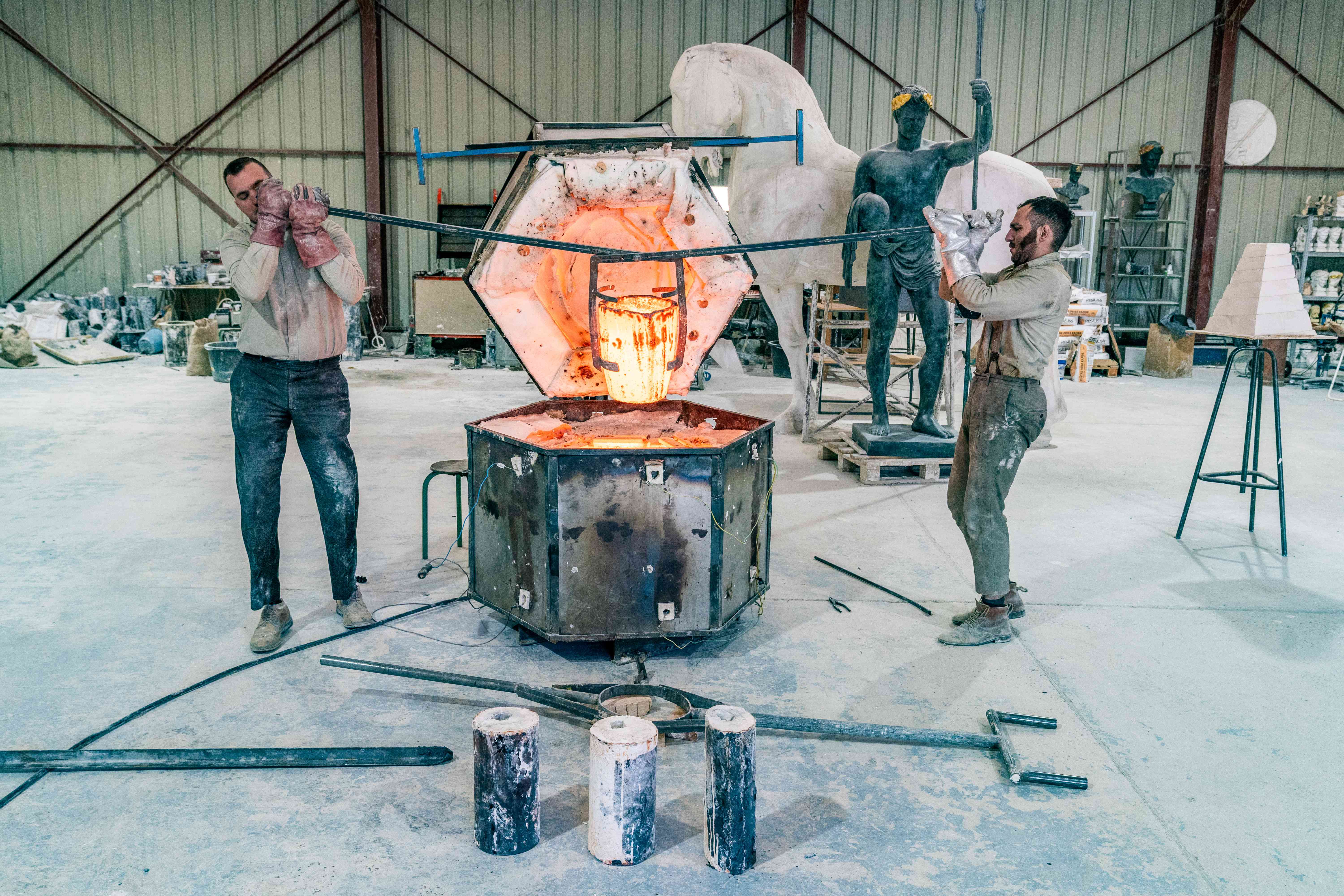 Deux artisants portent grâce à un outil un saut rempli de bronze fondu.